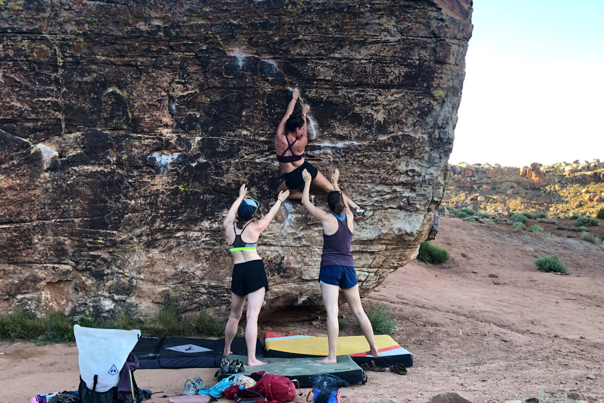 Two women spotting a third woman while she climbs a boulder over boulding crash pads