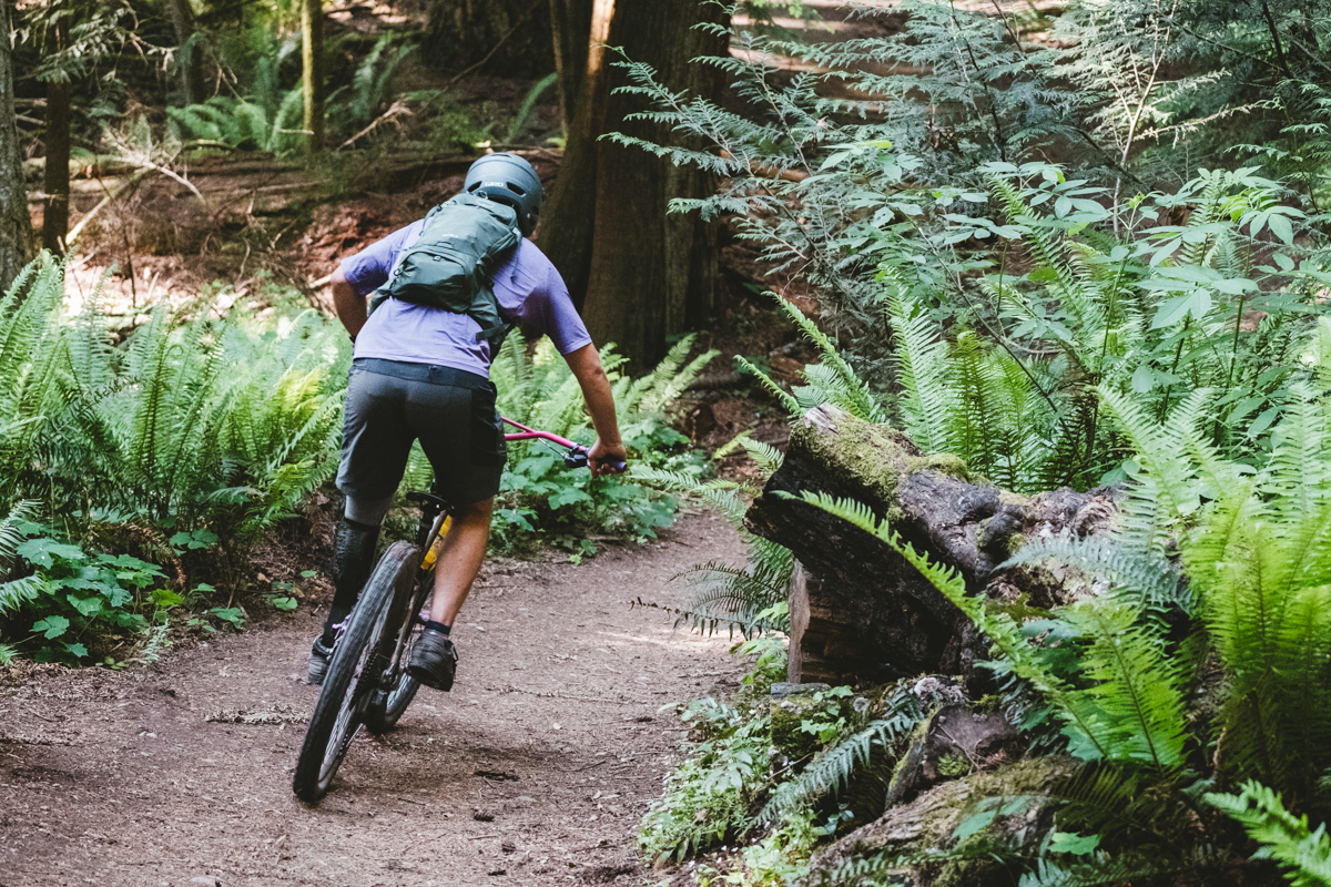 A man riding a bike down a forested trail while wearing a green mountain biking backpack