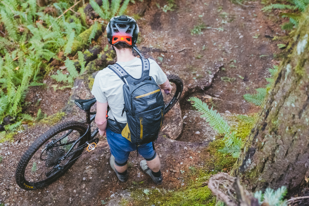 A man standing next to his mountain bike on a forested trail wearing a black mountain bike backpack