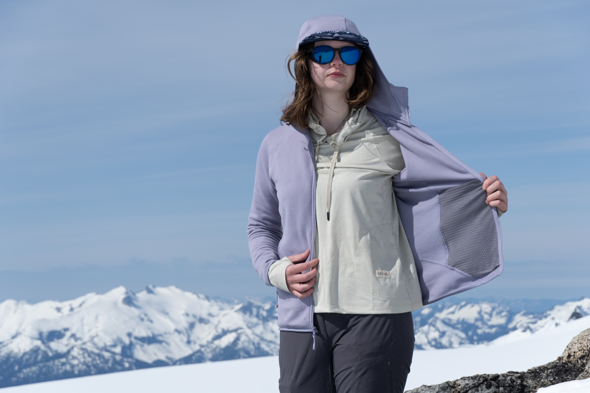 A woman putting on a fleece jacket before a snowy mountain background