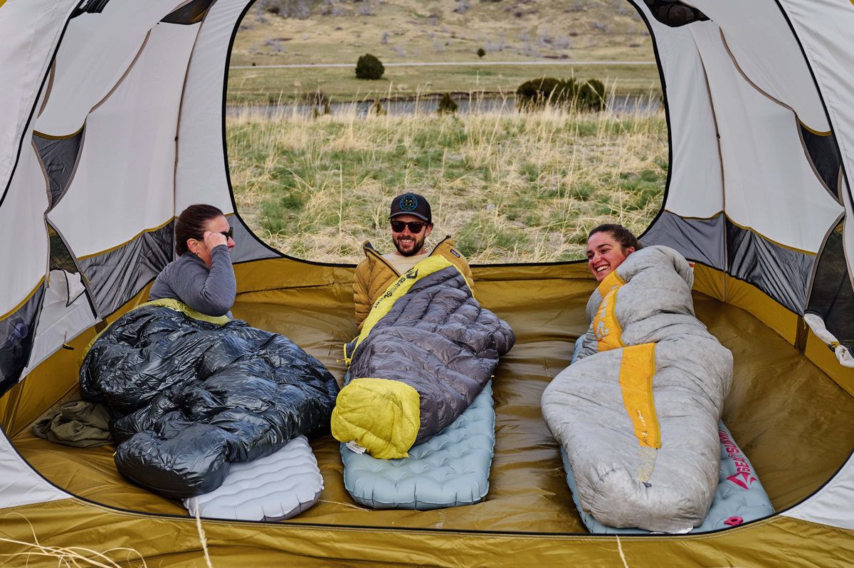 Three campers spread out inside a camping tent with a river background