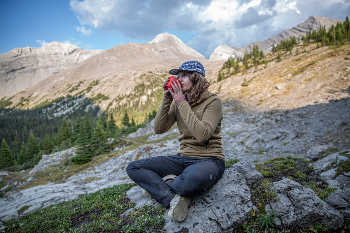 A woman drinking from a camp mug in the mountains wearing a brown fleece jacket