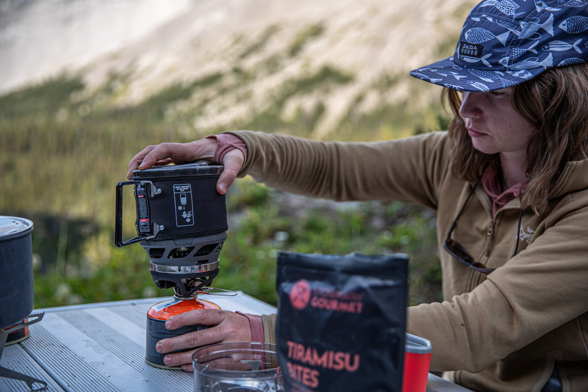 A woman fiddling with a backpacking stove while wearing a brown fleece jacket