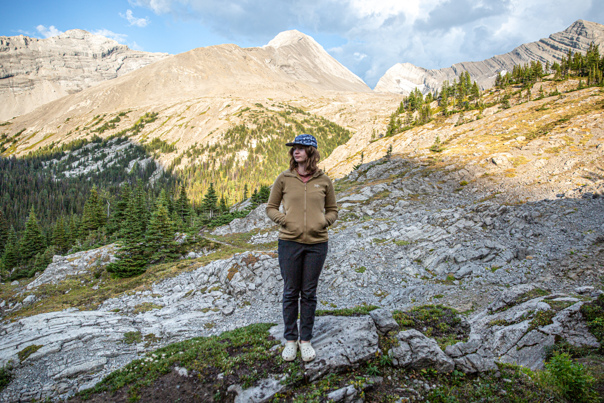 A woman standing in front of a mountainous landscape with her hands in the pockets of a brown fleece jacket