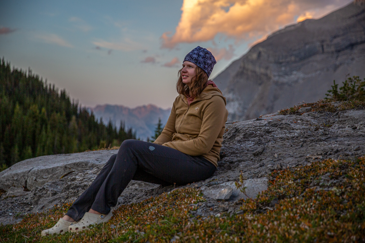 A woman stting on a rock in a mountainous landscape at sunset in a brown fleece jacket