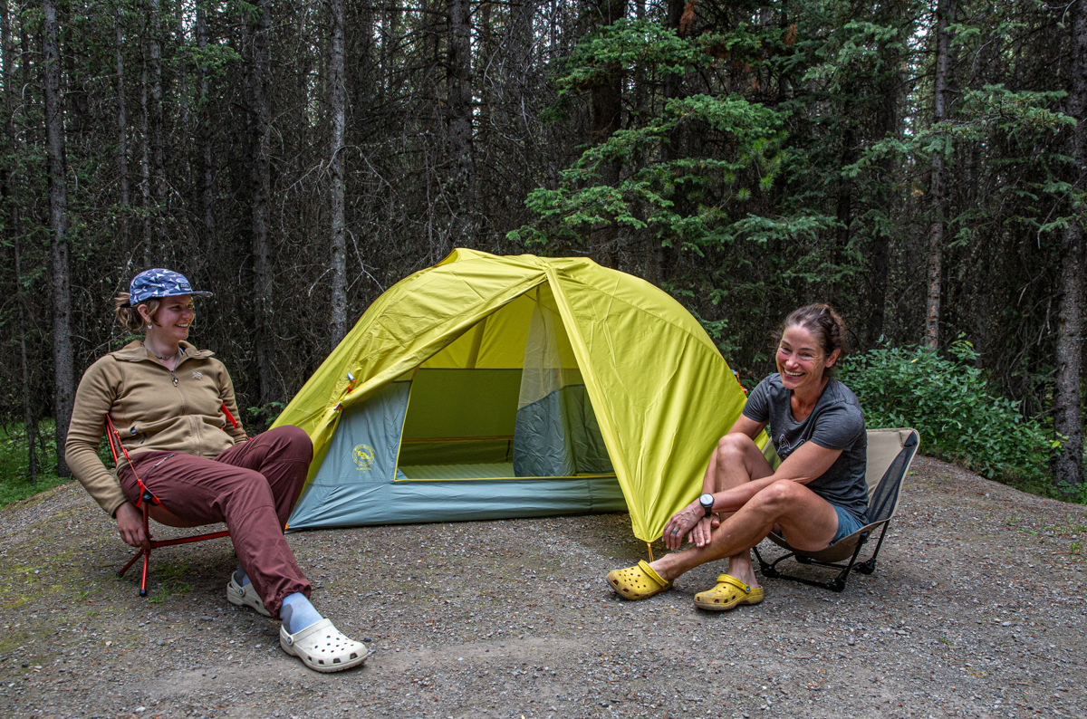 Two women sitting in chairs in front of a backpacking tents and wearing a brown fleece jacket