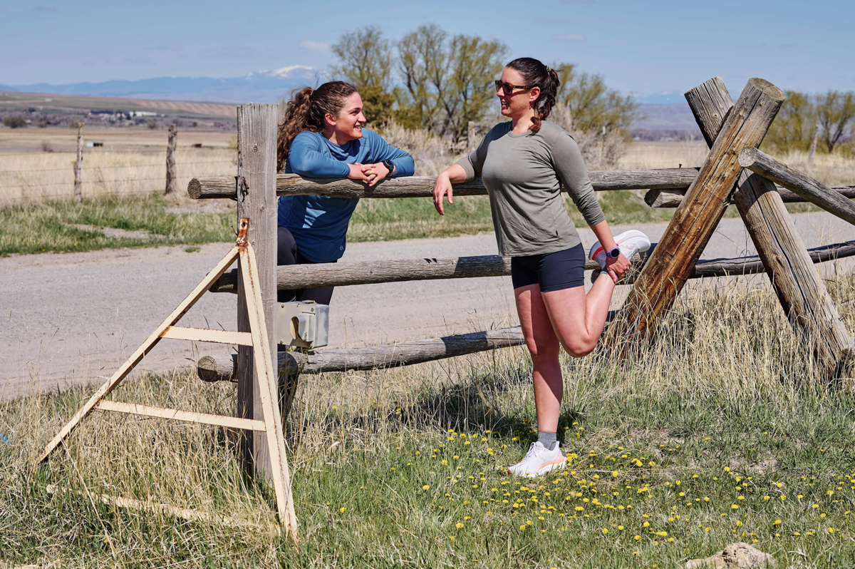 Two women leaning on wooden fence and streching legs while wearing purple fitness watch