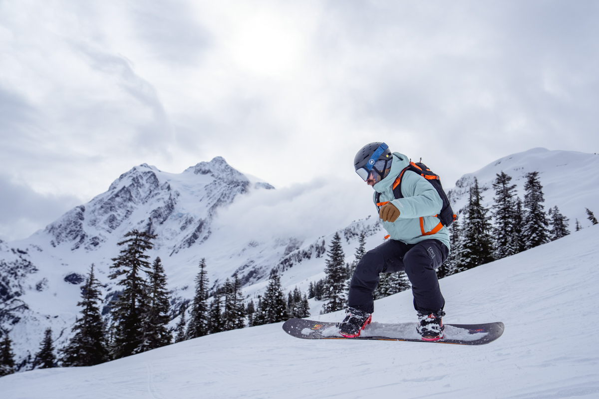 A man snowboarding over a snowy slope with a mountain background while wearing a red ski backpack