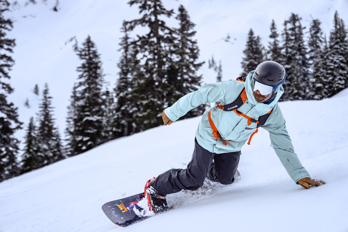 A man snowboarding down a slope touching the snow while wearing a red ski backpack
