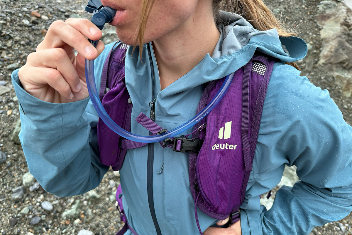 A close up of the Deuter logo on a purple daypack while a woman drinks out of a hydration hose