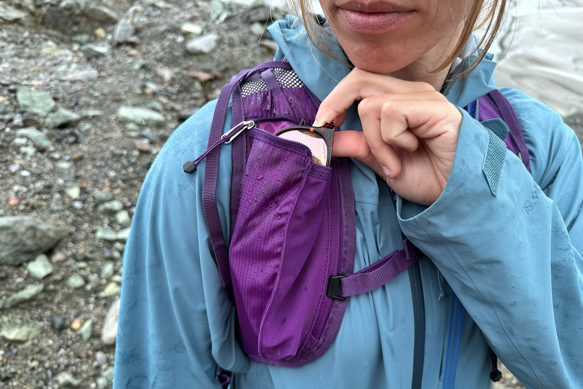A hand pulling out a pair of sunglasses out of a shoulder strap pocket on a purple daypack
