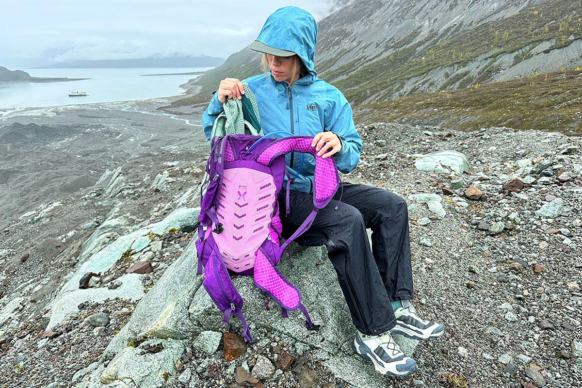A woman pulling a fleece jacket out of a purple daypack while sitting on a rock
