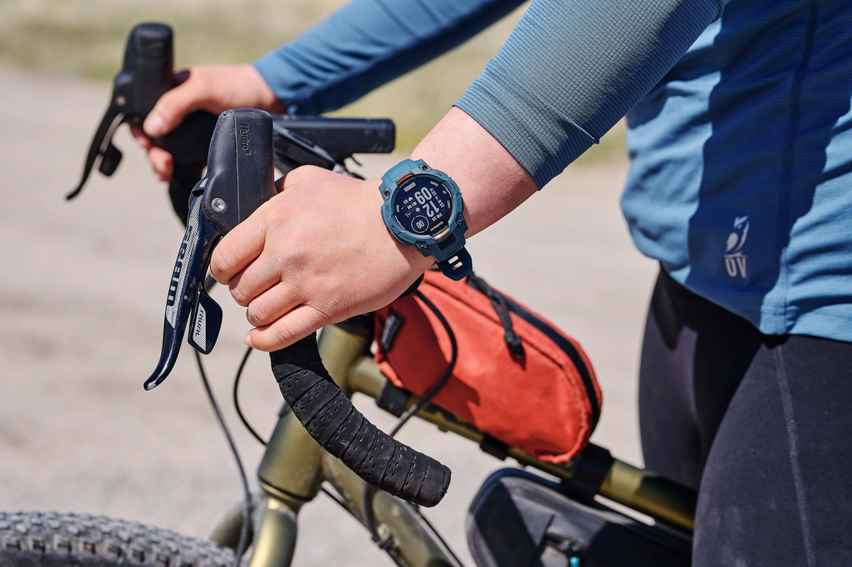 A close up of a blue GPS watch on a wrist with a hand holding bike handle bars