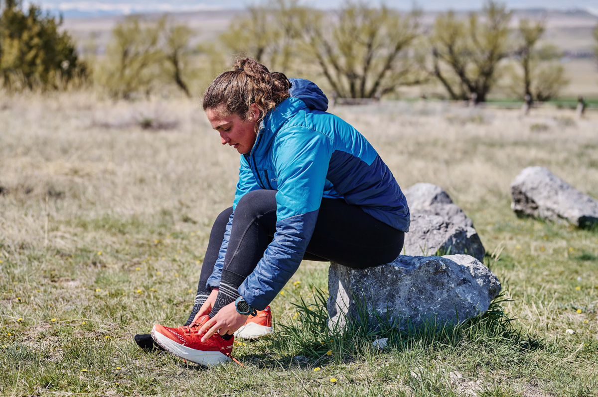A woman sitting on a rock and tying her running shoes while wearing a blue GPS watch