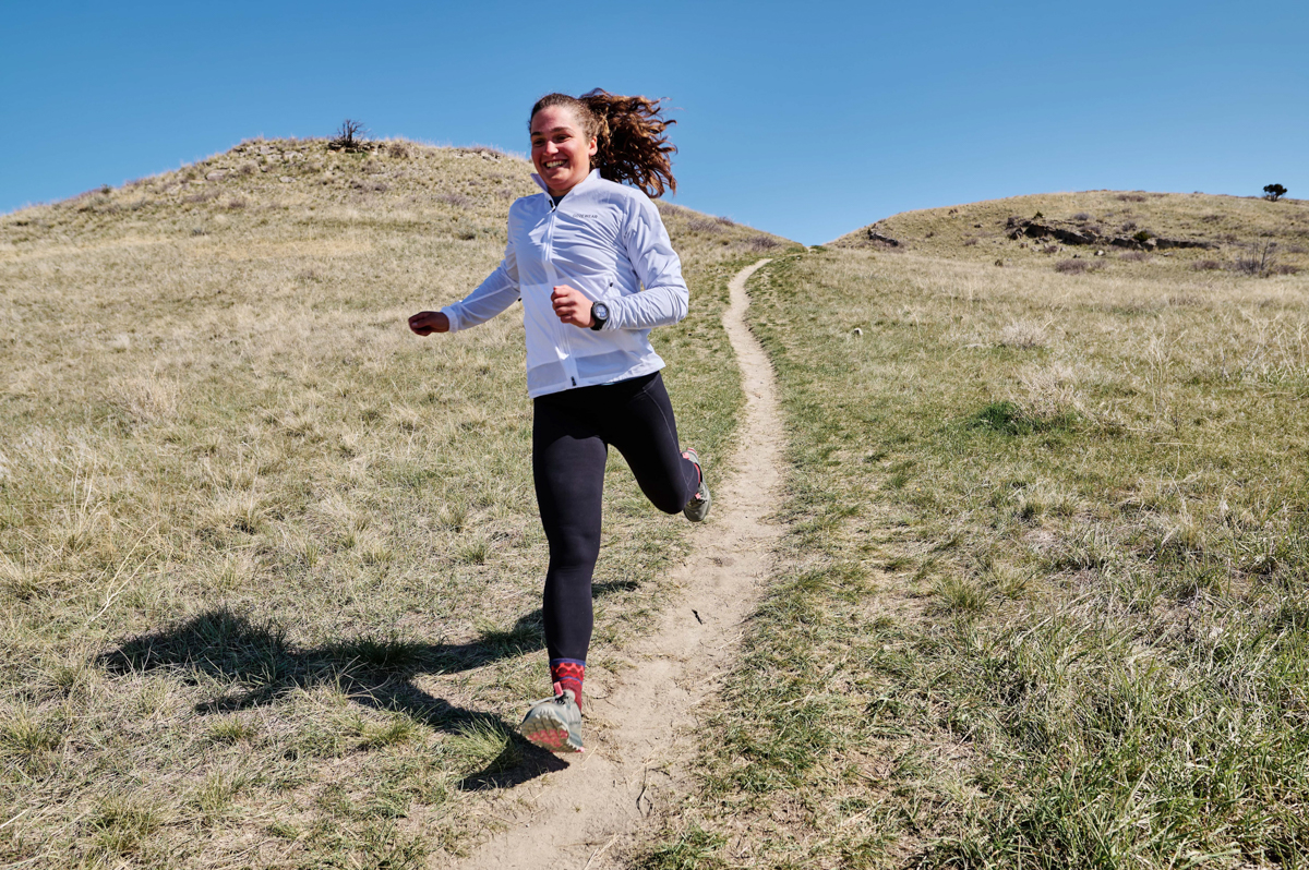 A woman smiles while running downhill on a grassy hill trail while wearing a blue GPS watch
