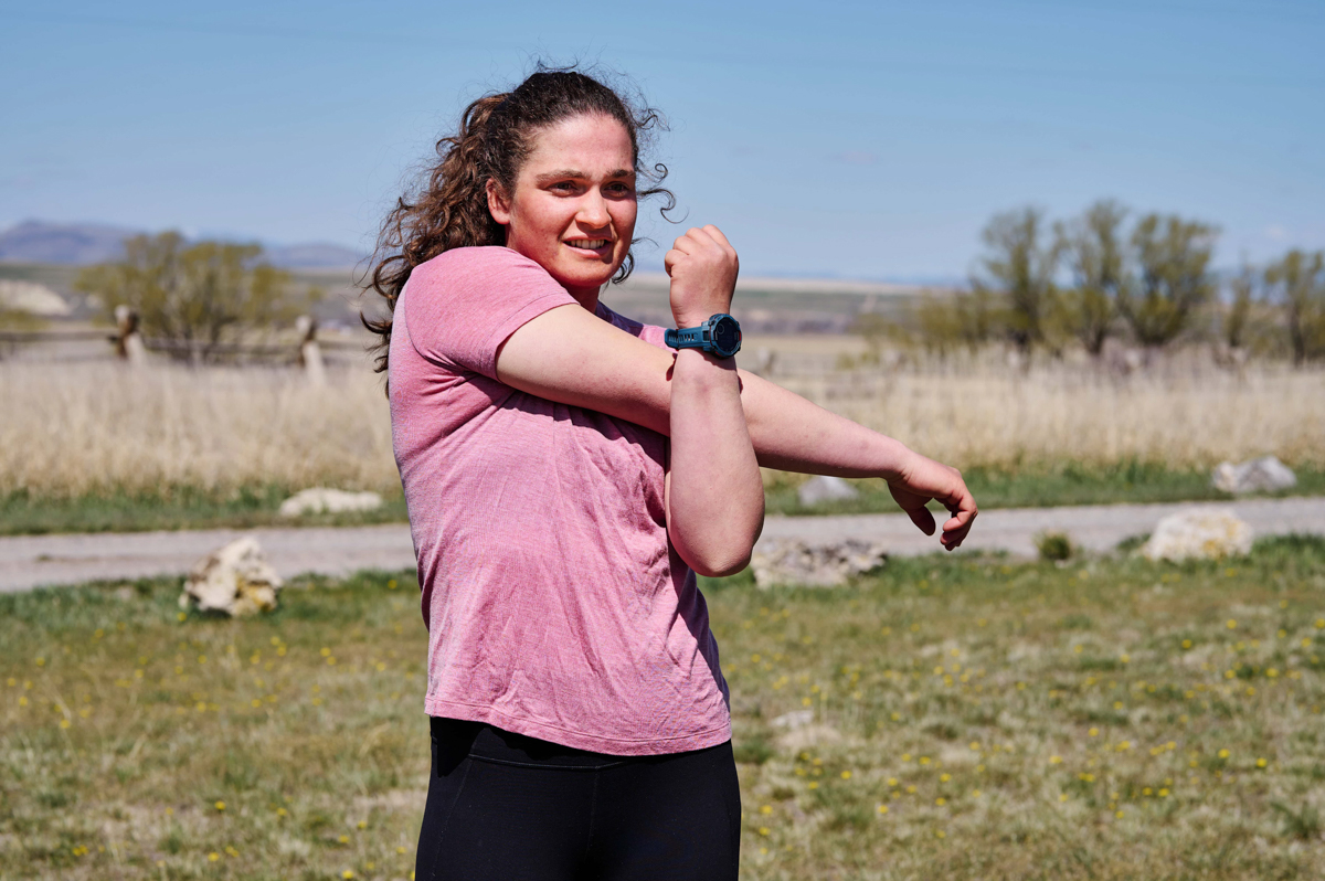 A woman stretching her arms in front of a grassy field while wearing a blue GPS watch