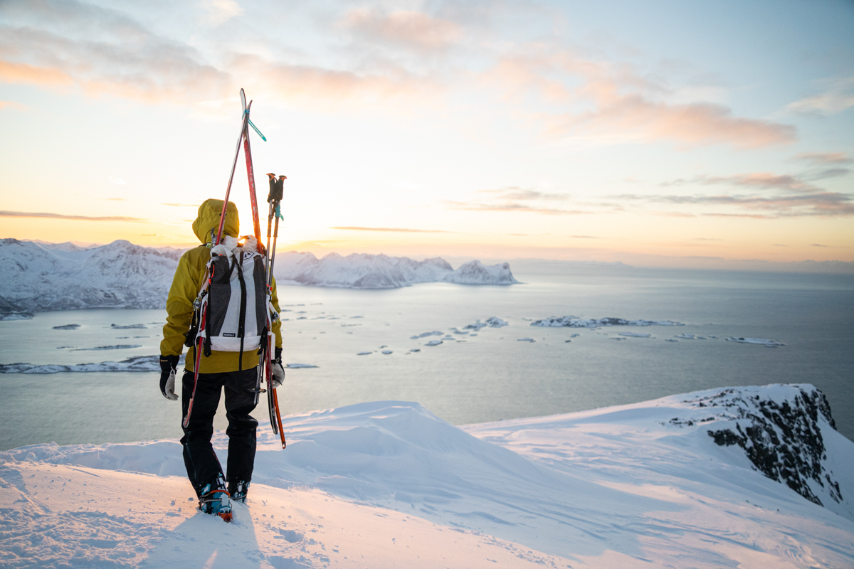 A man in a yellow jacket hiking on a snowy landscape with skis tied to his white ski backpack
