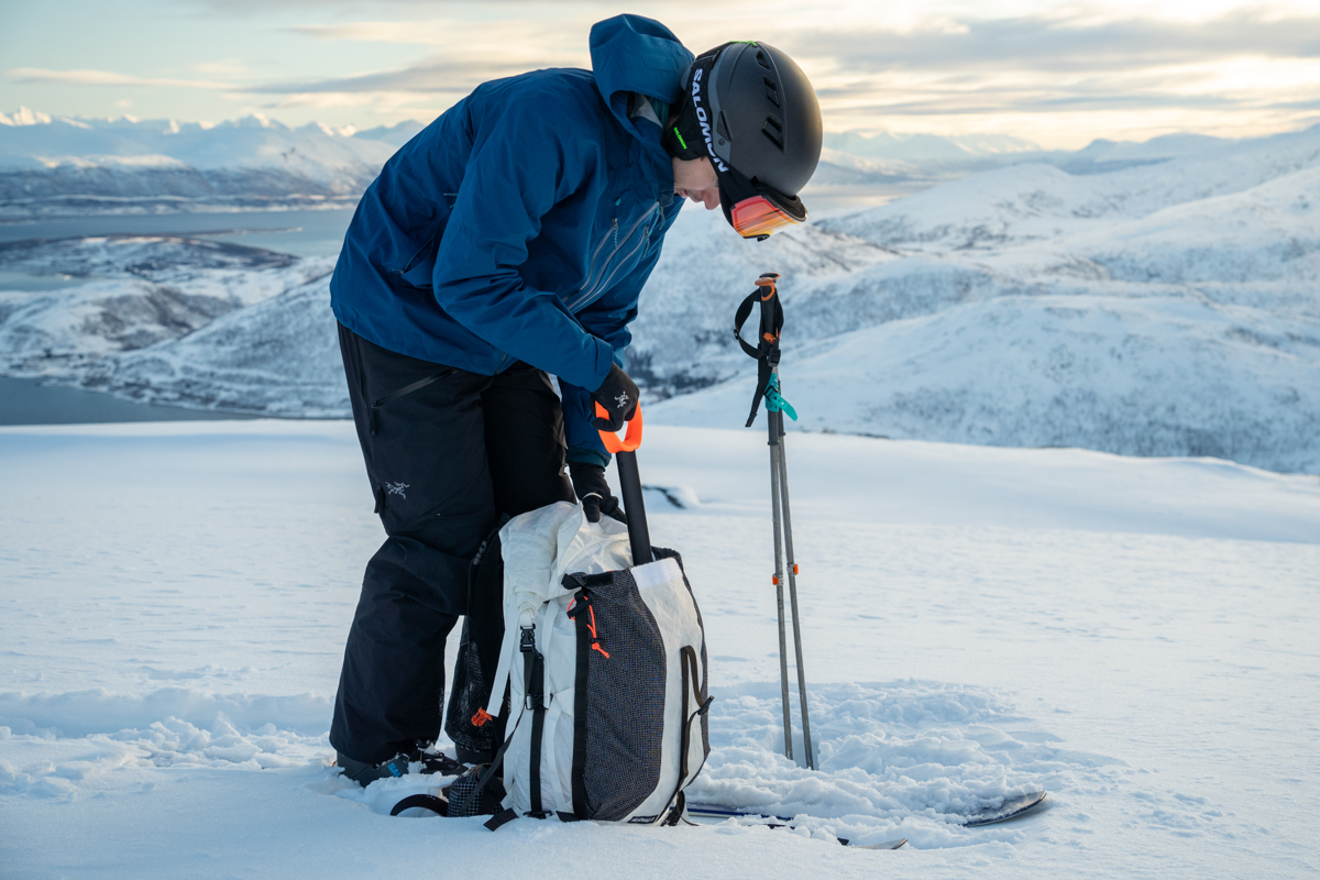 A man pulling a snow shovel out of a white ski backpack in a snowy landscape