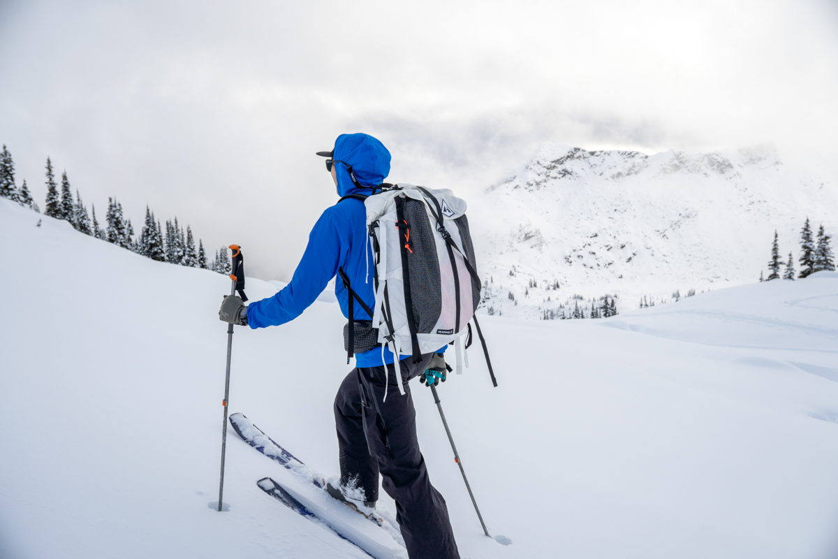 A man ski touring up a snowy hill wearing a blue jacket and white ski backpack