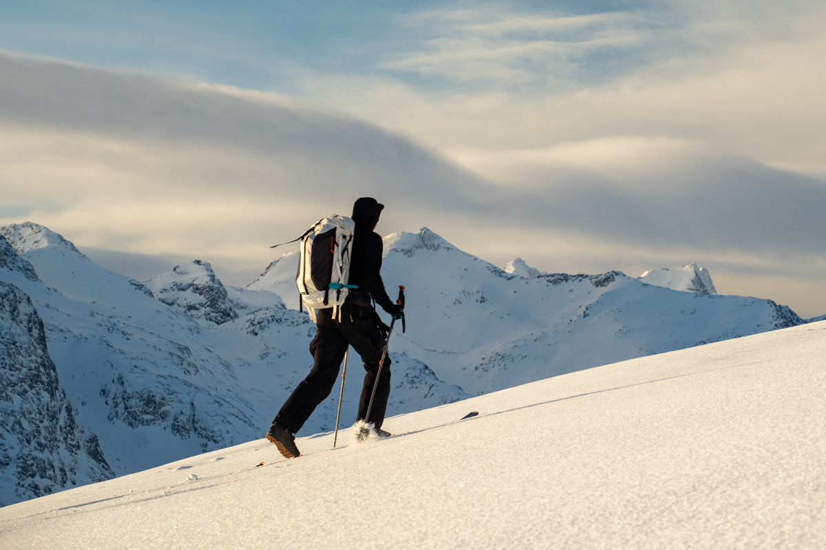 A man ski touring up a snowy slope in a mountainous landscape wearing a white ski backpack
