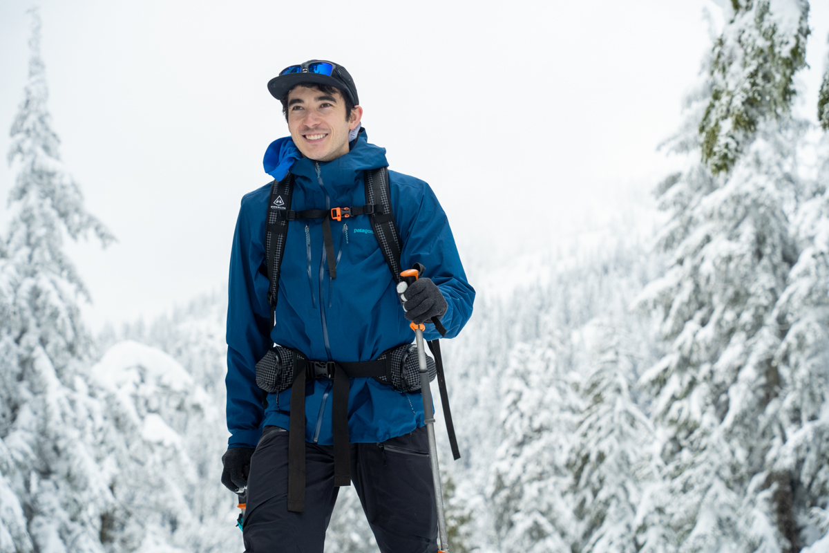 A man smiling while skiing in a blue jacket and white ski backpack