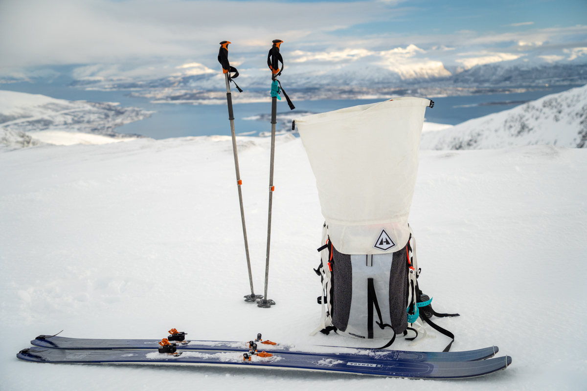A white ski backpacking sitting in the snow beside a pair of ski poles and skis in a mountainous landscape