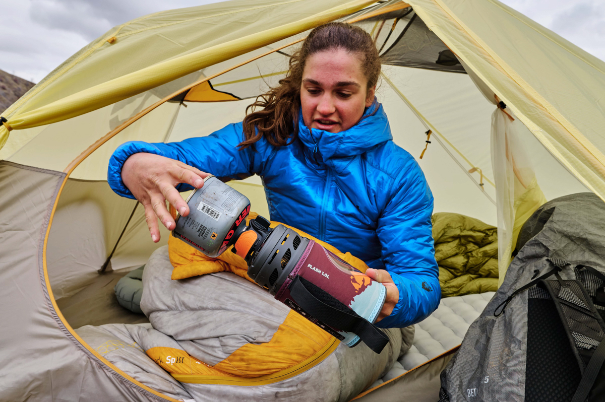 A woman in a tent screwing a fuel canister onto the bottom of a backpacking stove
