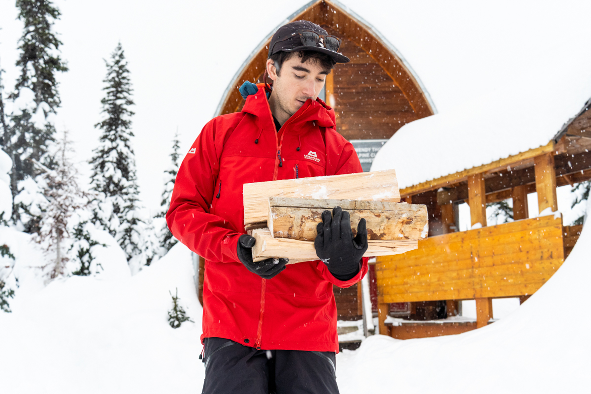 A man carrying split wood in front of a snowy cabin in a red hardshell jacket