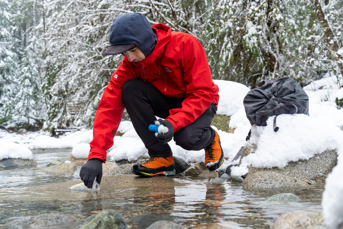 A man gathering water from a snowy stream in a red hardshell jacket