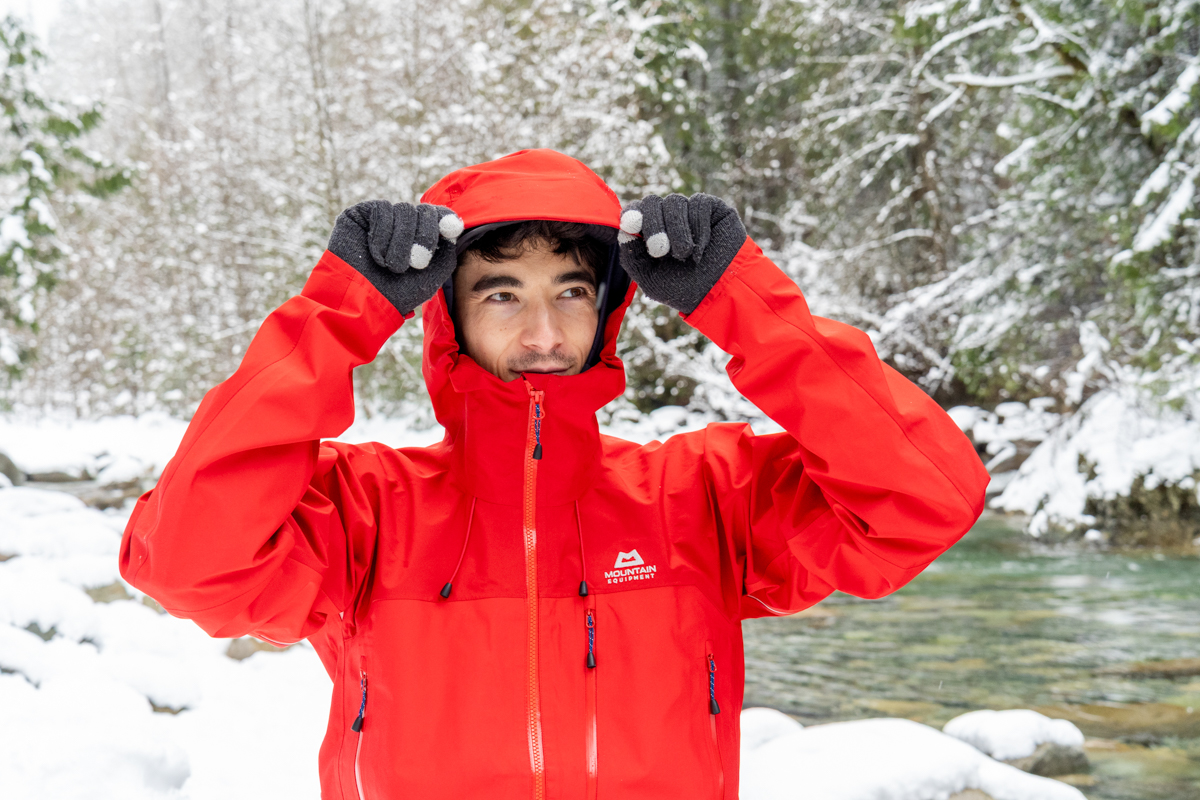 A man pulling the hood up on a red hardshell jacket with a snowy landscape