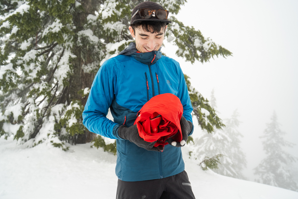 A man smiling as he rolls a red hardshell jacket into its hood in a snowy forest landscape