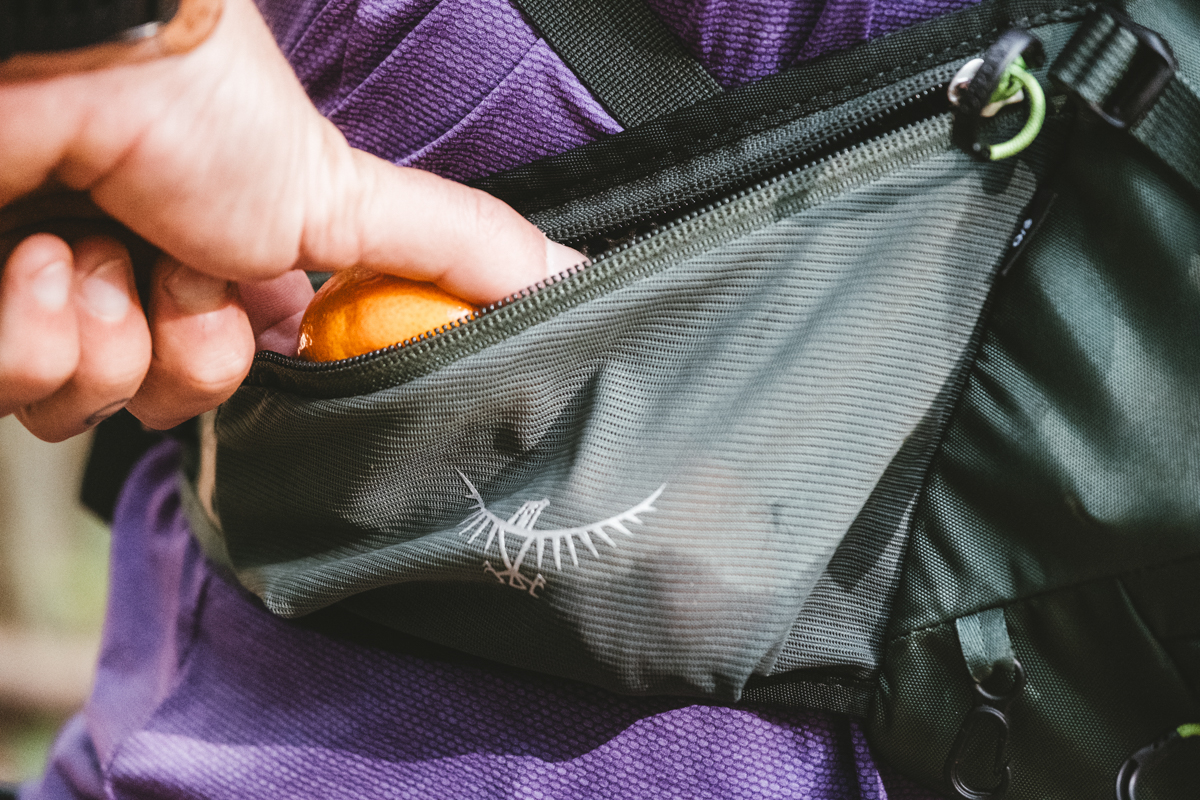 A close up of a hand pulling an orange out of a hipbelt pocket on a mountain bike backpack