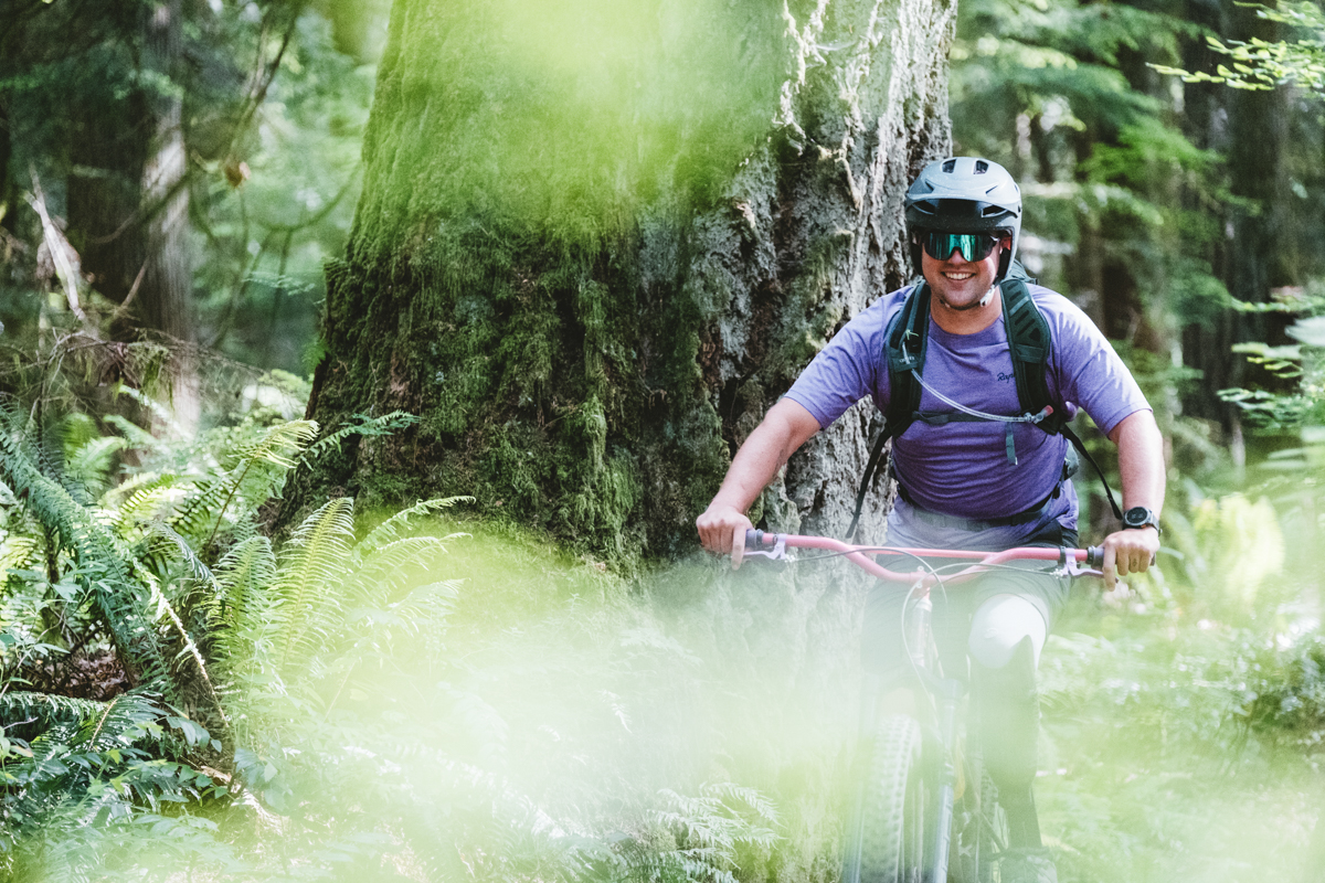 A man smiling while riding a bike through a forest and wearing a mountain bike backpack