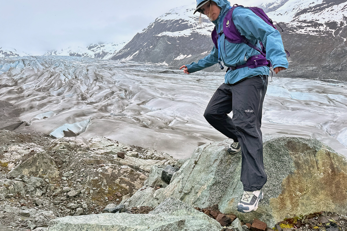 A woman stepping down onto a rock while hiking in a rocky and glaciated landscape in hiking shoes