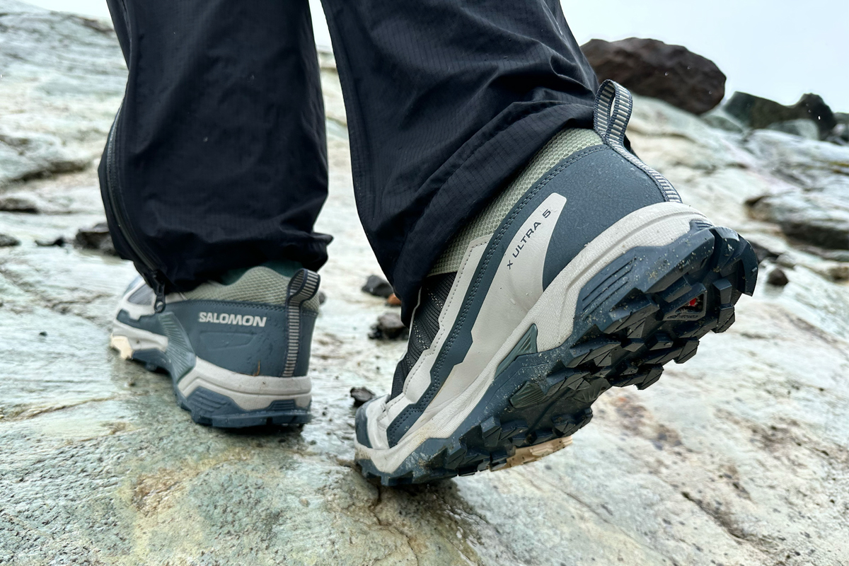 The green and brown Salomon X Ultra 5 hiking shoe close up and walking away across a rock
