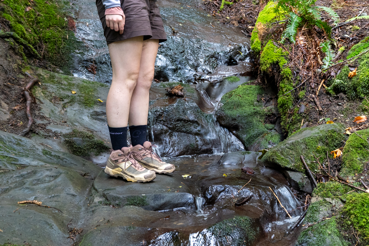 A close up of brown and pink hiking boots next to a small waterfall in a forest