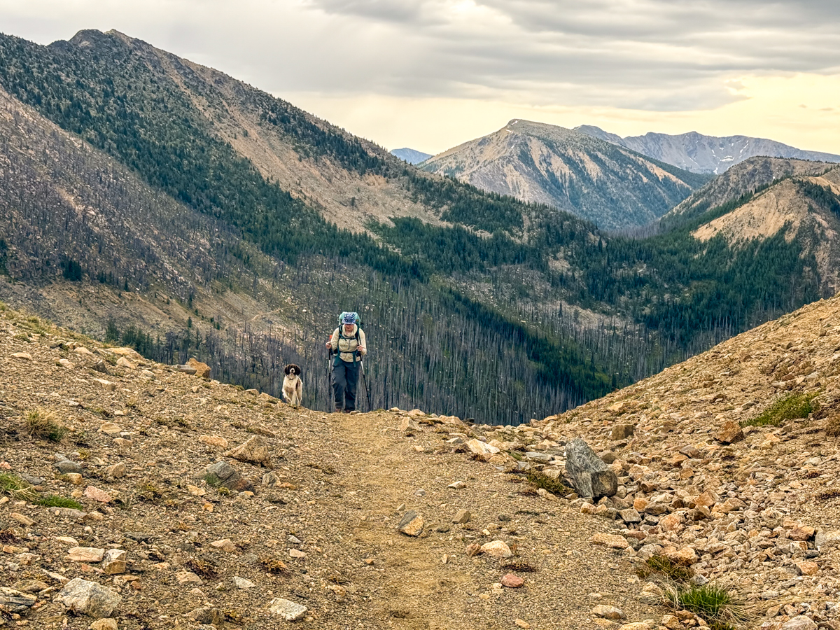A far away shot of a woman hiking in hiking boots up a steep dirt hill with a dog and mountains in the background