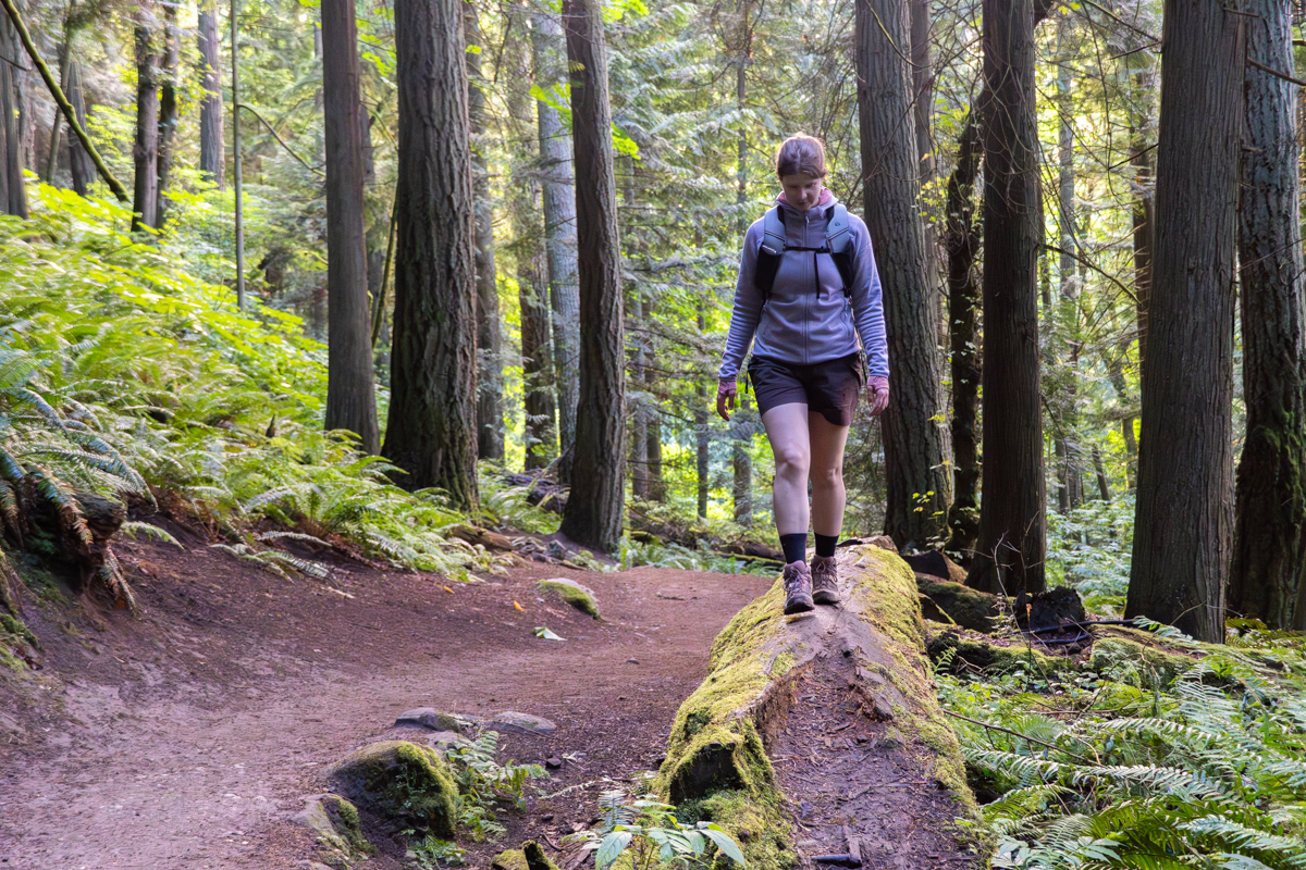 A woman walking on a log next to a trail in a forest wearing hiking boots