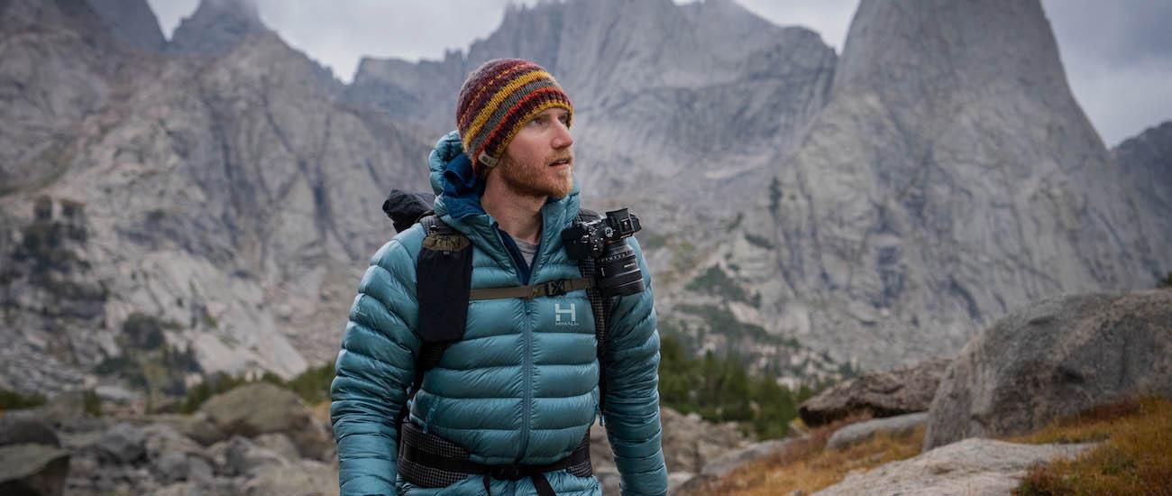 A man walks in the mountains carrying a backpack with a camera clipped to the shoulder strap