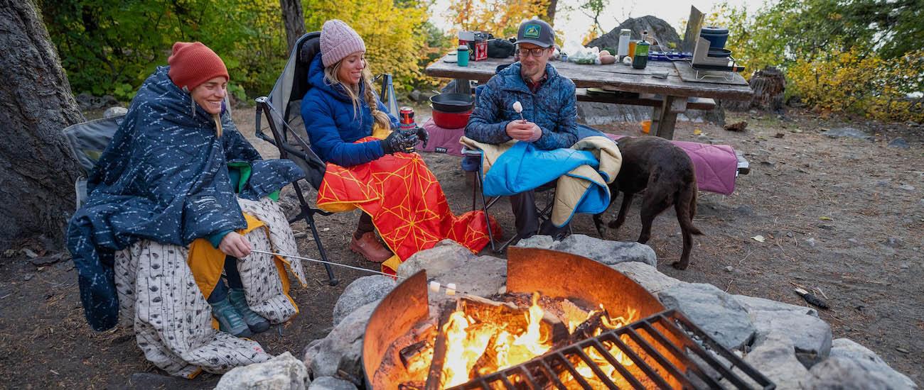 Three friends sit around a campfire under blankets while roasting marshmallows