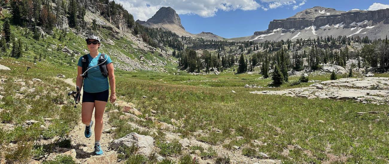 A woman wearing a running vest walks along a trail with mountains in the background