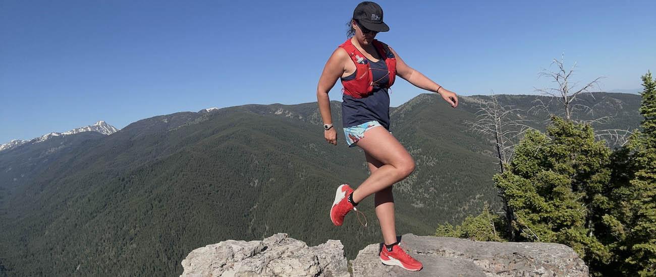 A woman shows off her red trail running shoes on top of a rock with mountains in the background