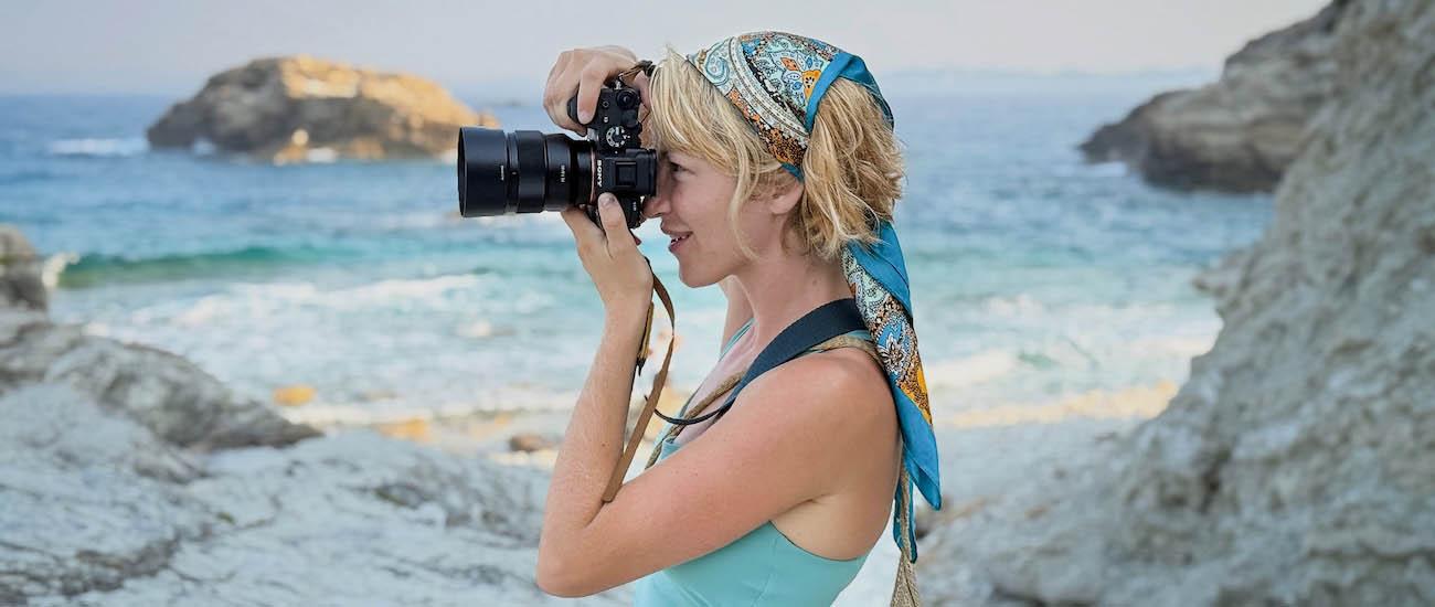 A woman standing next to the ocean looks through a camera lens