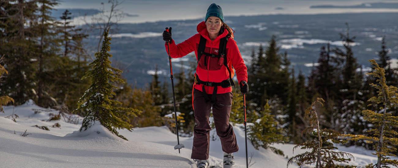 A woman in a red jacket skins up a hill on hill on skis with ski poles