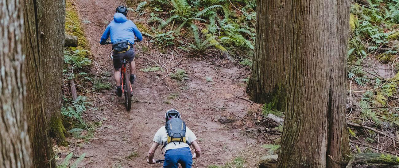Two bikers ride up a mountain bike trail wearing backpacks
