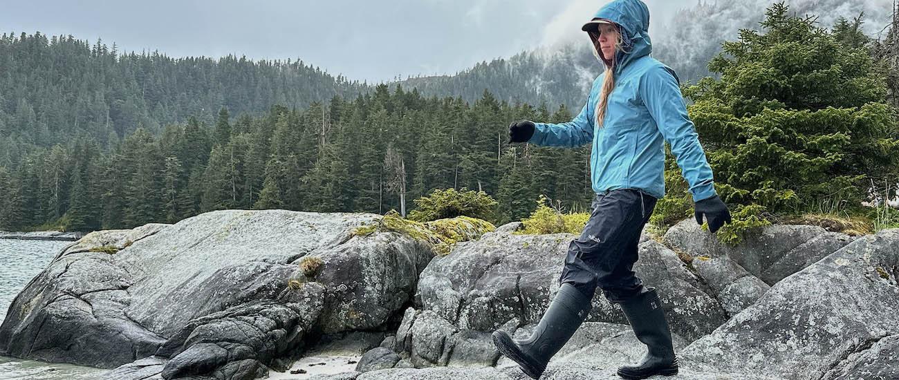 A woman in rain boots and rain jacket walks across rocks on a rainy day