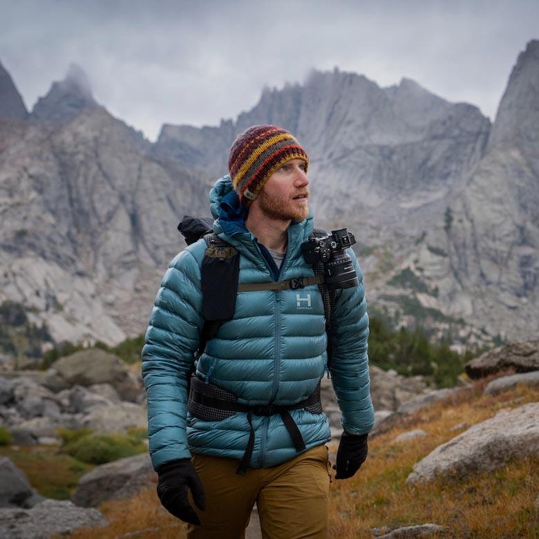 A man walks in the mountains carrying a backpack with a camera clipped to the shoulder strap