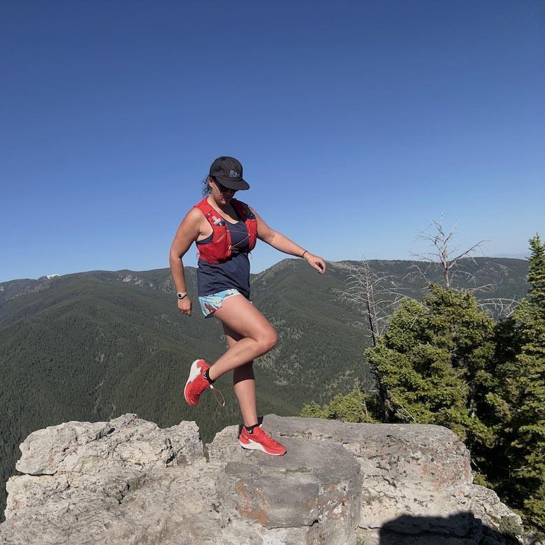 A woman shows off her red trail running shoes on top of a rock with mountains in the background
