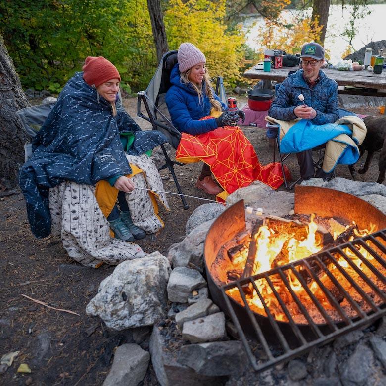 Three friends sit around a campfire under blankets while roasting marshmallows
