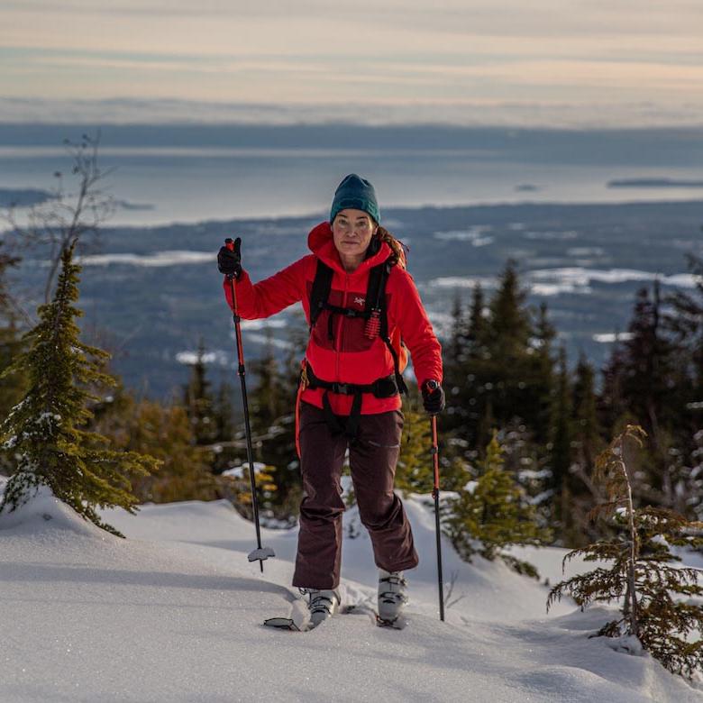 A woman in a red jacket skins up a hill on hill on skis with ski poles